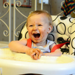 Jak naučit dítě jíst lžičkou, smějící se dítě u stolečku, špinavé od jídla